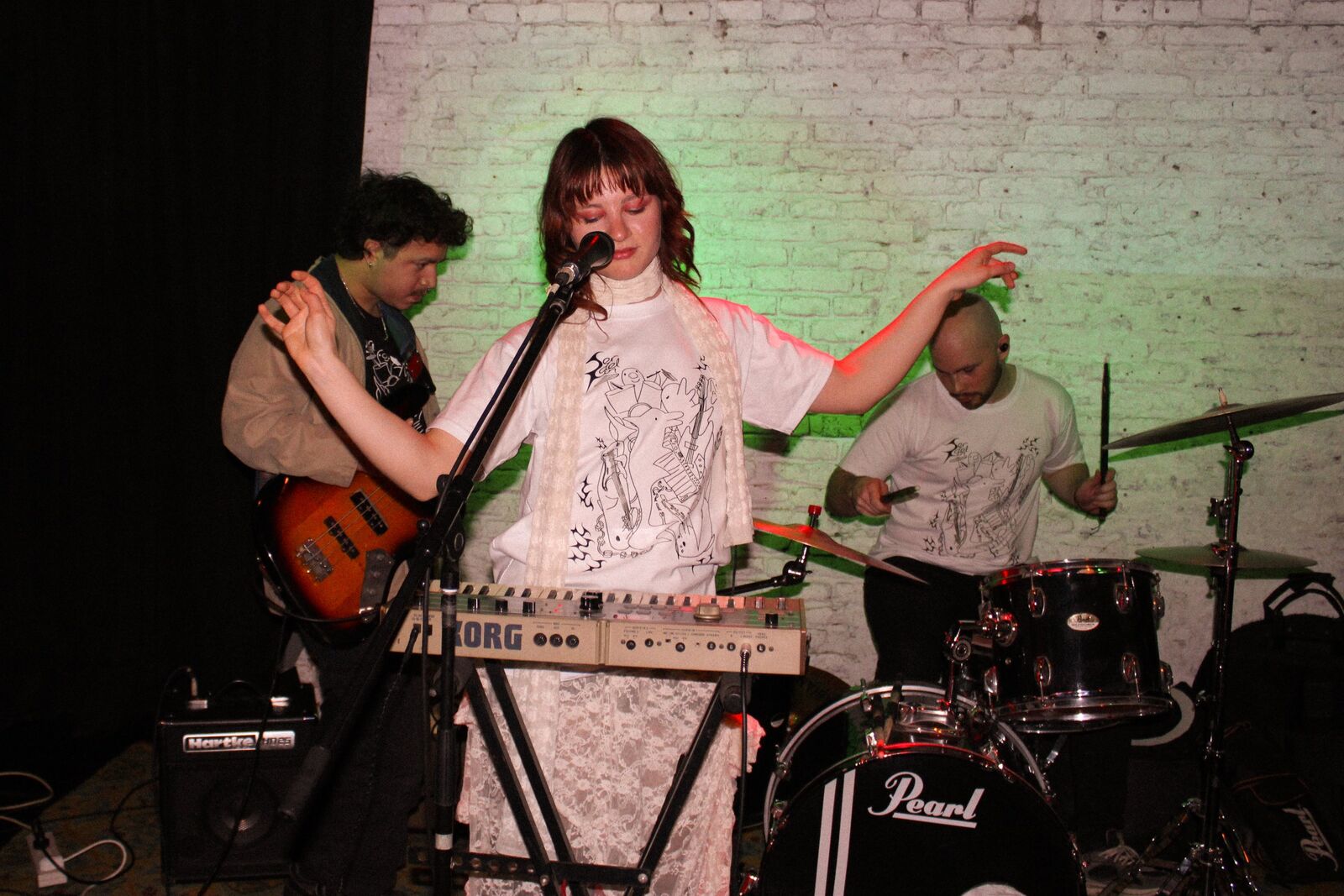 Cantante con los brazos abiertos detrás de un teclado, con bajista y baterista atrás bajo luz verde