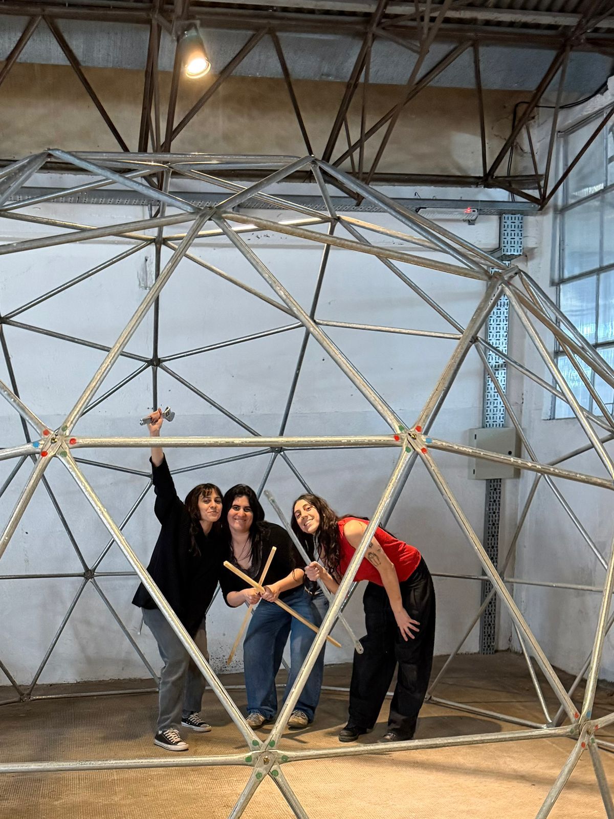 Las tres artistas sonrientes dentro de la estructura del domo, una con una llave en alto y otra con dos varillas de madera cruzadas