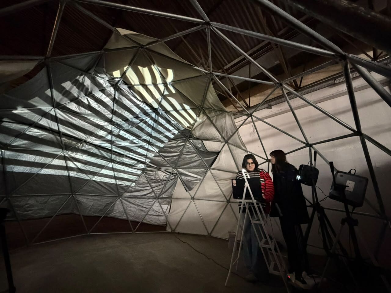 Interior del domo con la estructura metálica cubierta de tela y una proyección de franjas de luz; dos de las artistas trabajan en una computadora junto a los proyectores