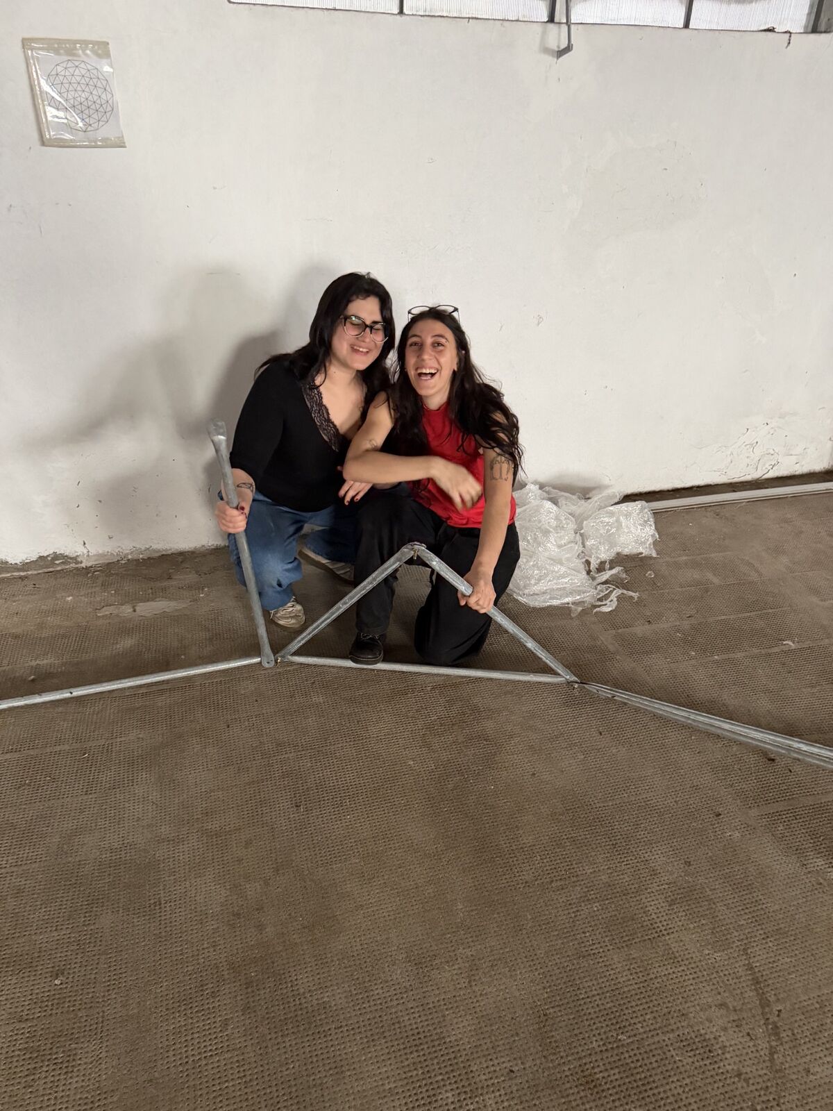 Dos de las artistas riéndose, agachadas junto a un triángulo de caños en el piso