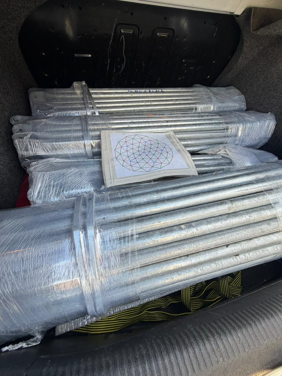 Caños galvanizados del domo envueltos en film dentro del baúl de un auto, con el plano de la estructura encima