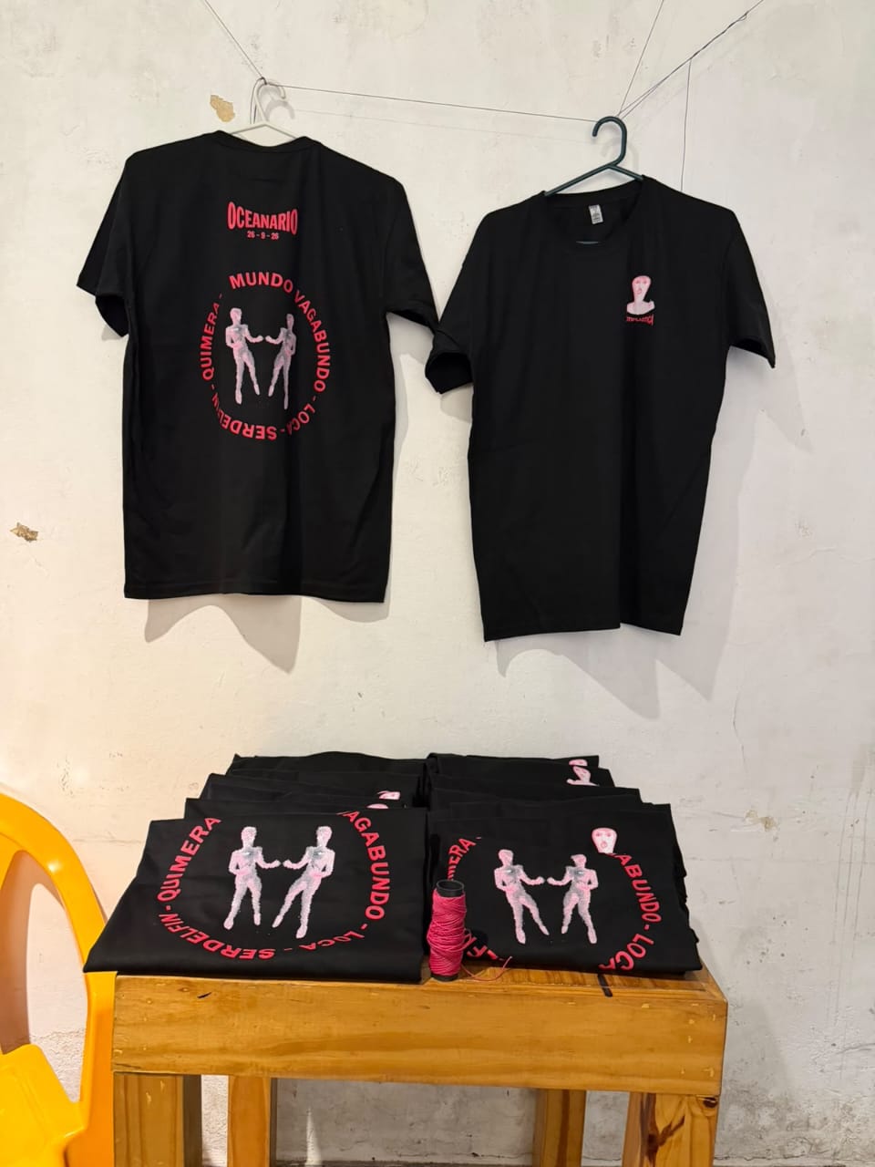 Dos remeras negras colgadas en la pared, una de espalda y otra de frente, y más remeras dobladas sobre una mesa
